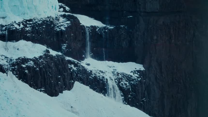 Meltwater from a glacier cascades down a snow-covered, rocky mountain cliff face