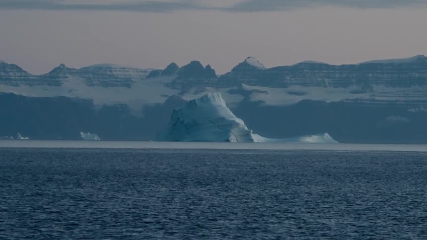 A massive iceberg floats majestically in the calm arctic ocean with a snow-covered mountain range in the background at dusk