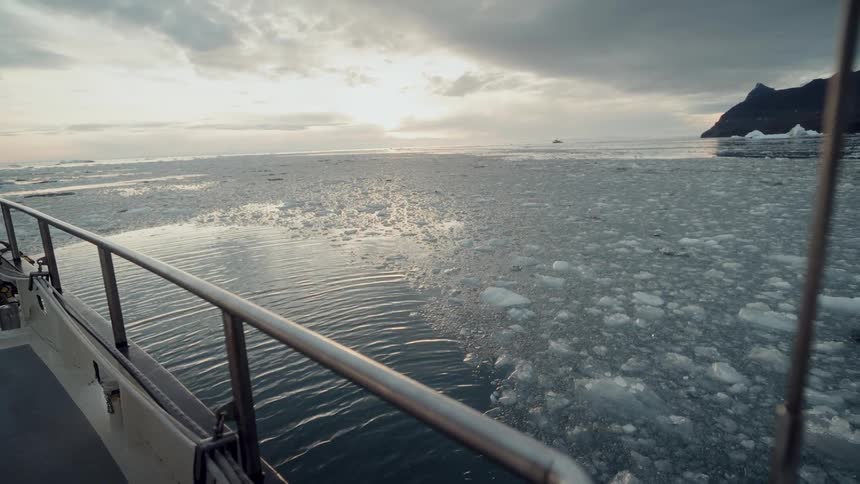 A boat glides through a tranquil, ice-filled sea as the sun sets under a dramatic, cloudy sky