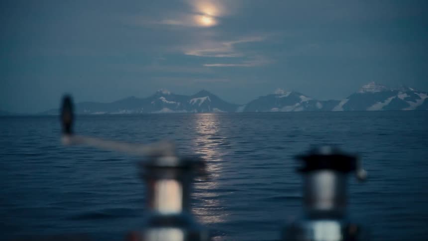 A serene view from a sailboat captures the moon reflecting on the tranquil sea with majestic snow-capped mountains in the distance at night