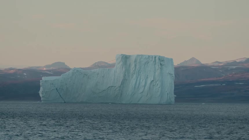A massive iceberg floats peacefully in the Arctic ocean at dusk with distant, snow-capped mountains in the background