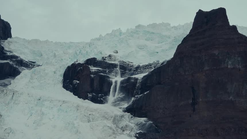 A powerful waterfall cascades down from a massive glacier on a rugged, rocky mountain under an overcast sky
