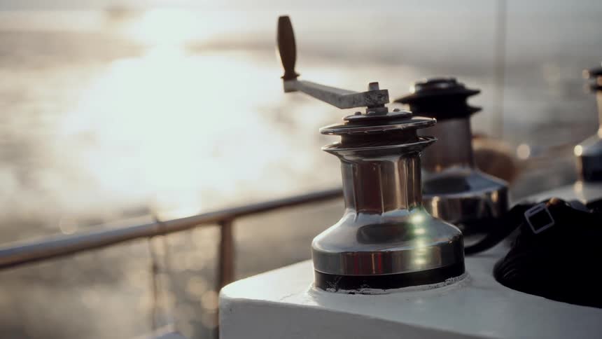 A close-up view of a sailboat's chrome winch and handle glimmers in the golden light of sunset while sailing on calm waters