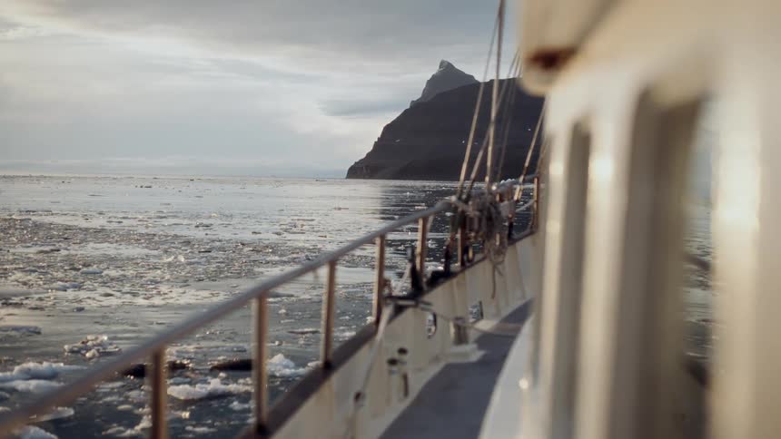 A boat navigates through a serene, ice-filled arctic sea at sunset, with a majestic mountain in the distance