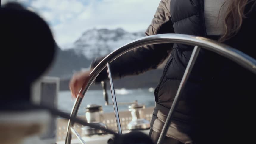 A close-up of a woman's hand steering a sailboat through a majestic, snowy mountain landscape