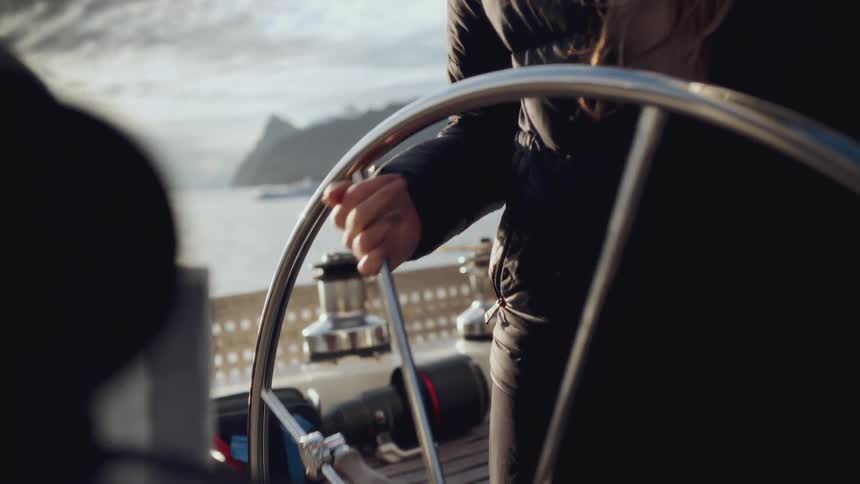 A close-up of a woman's hands steering a sailboat's chrome helm along a mountainous coast in the soft morning light