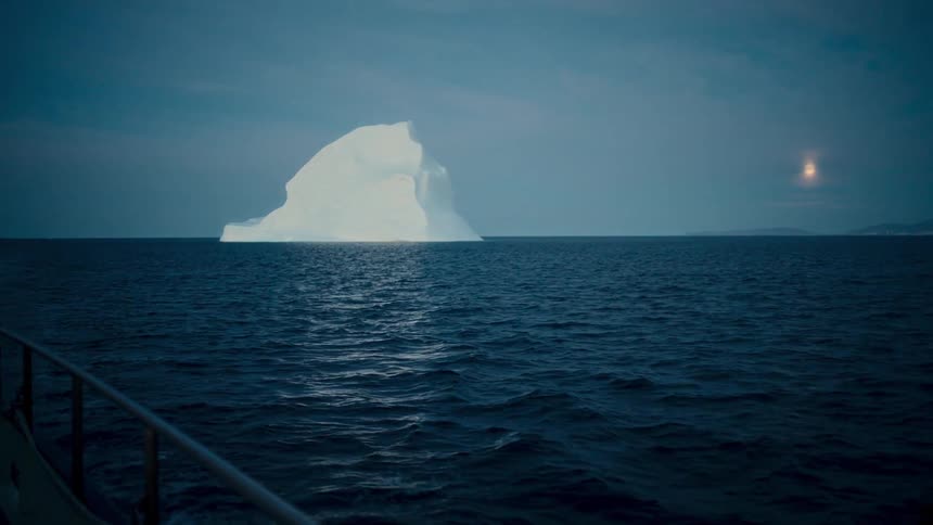 View from a boat of a large iceberg floating in the deep blue ocean at night with a full moon in the sky