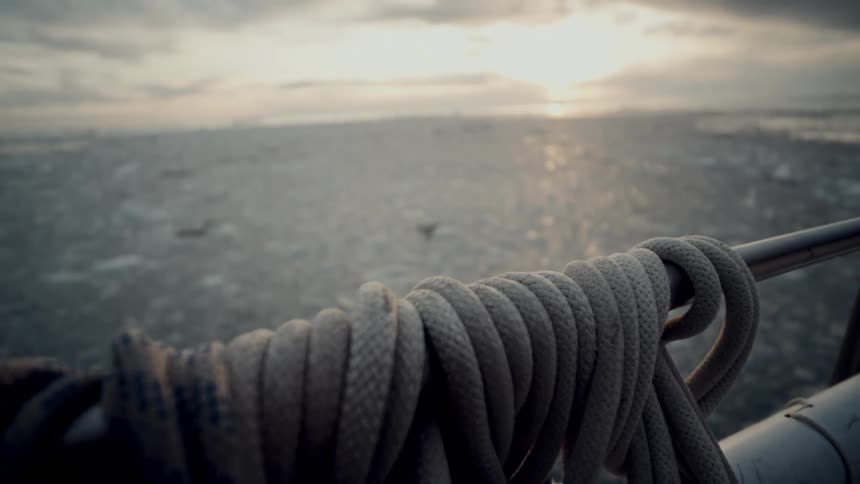 A dramatic rack focus from a coiled rope on a boat rail to a vast, icy sea at sunset