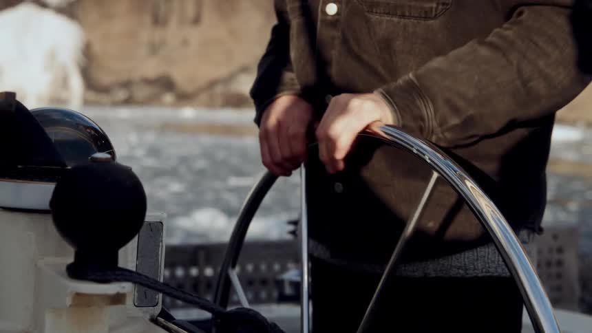 A close up shot of a man's hands on the helm, steering a boat through a frozen river in the winter