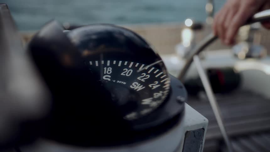 Close-up of a sailboat's binnacle compass pointing southwest as a person steers the helm on the water