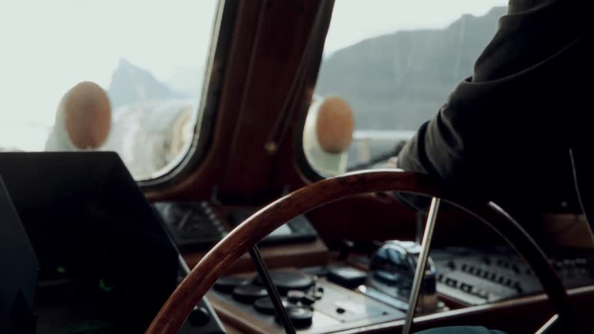 A captain pilots a vintage wooden boat through a cold, mountainous landscape as seen from the wheelhouse