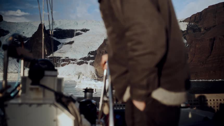 An explorer on a sailboat gazes in awe at a majestic glacier cascading down a mountain into the arctic sea