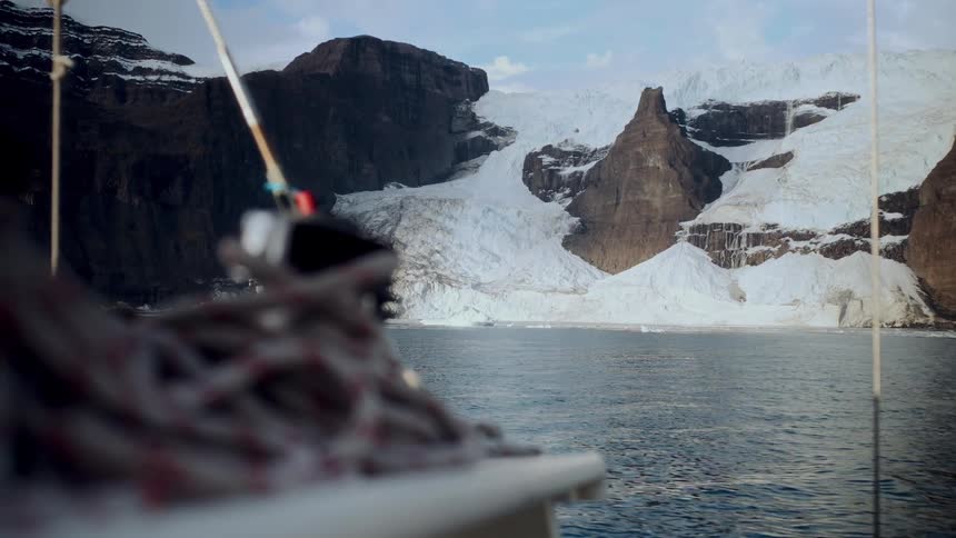 A sailboat glides through calm arctic waters, approaching a massive glacier nestled in the rugged mountains