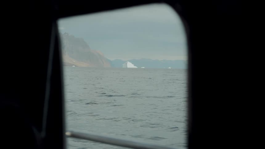 Through a dark boat window, a distant iceberg and mountains are visible across the vast, cold ocean