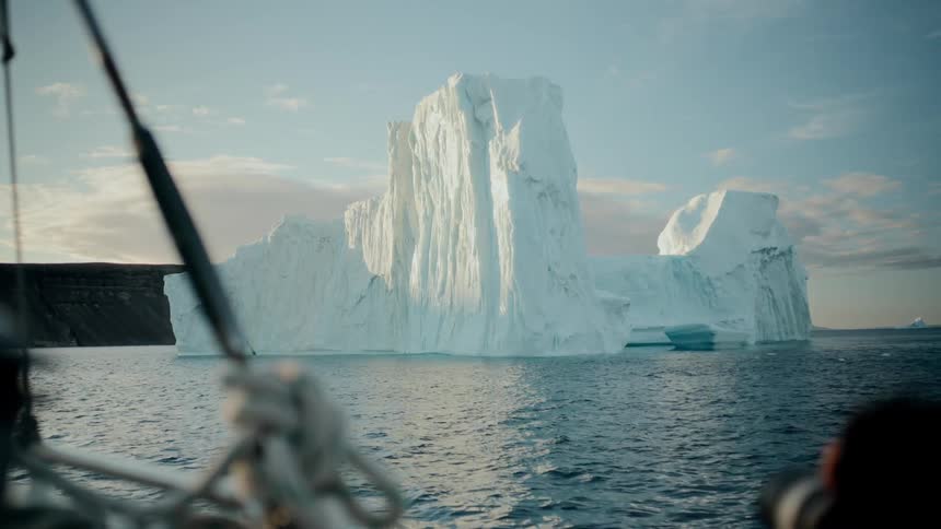 A boat sails slowly past a massive, towering iceberg floating in the calm Arctic Ocean at sunrise
