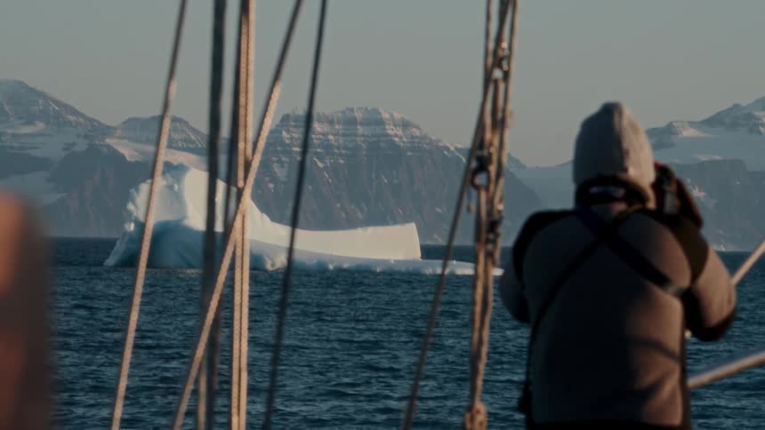From the deck of a sailboat, a traveler observes a large iceberg adrift in the sea with snow-covered mountains on the horizon
