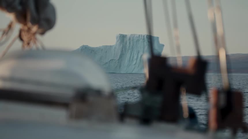 A massive iceberg floats in the calm ocean, viewed from the out-of-focus deck of a passing sailboat