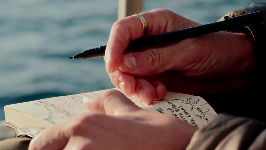 Close-up of an artist's hands sketching and journaling in a notebook with the calm sea in the background
