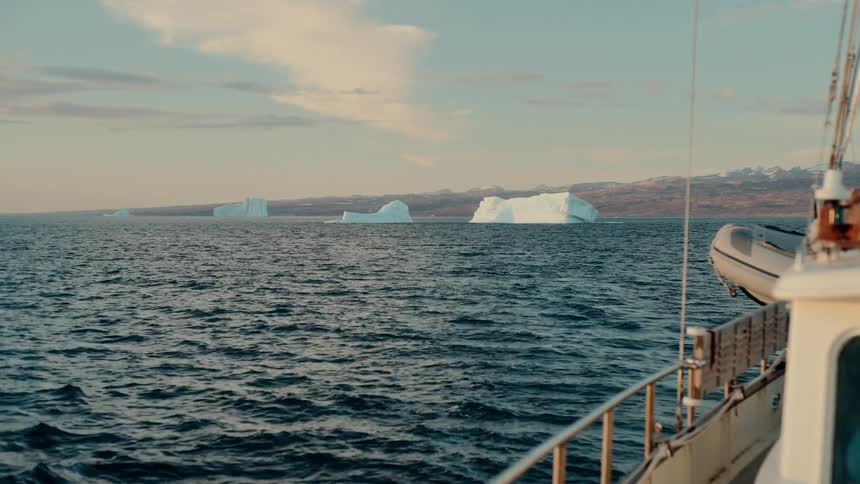 View from a boat sailing across the arctic ocean towards massive icebergs along the distant coastline at sunset