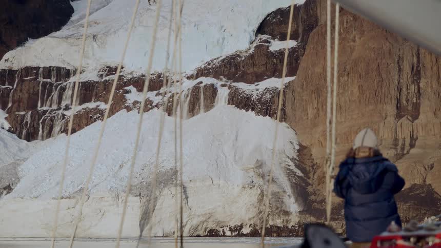 A person in winter gear stands on the deck of a boat, observing a powerful waterfall on a massive, snow-covered mountain