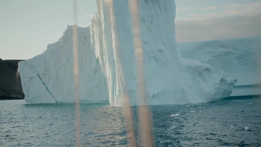 A stunning slow reveal from behind a silhouette shows a massive iceberg with melting waterfalls, as seen from a moving boat in the Arctic Ocean