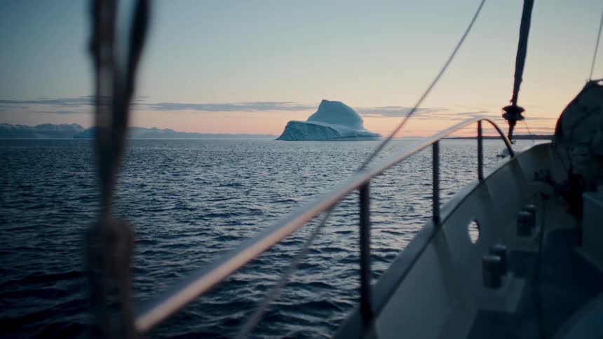 A view from a sailboat showing a massive iceberg floating on the calm ocean at sunset, with distant mountains on the horizon