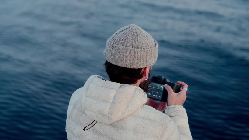 A male photographer in a warm jacket and beanie captures the serene blue ocean with his camera in slow motion