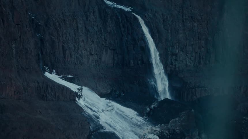 A powerful waterfall carves a path down a dark, rugged cliff face, meeting a field of snow at its base
