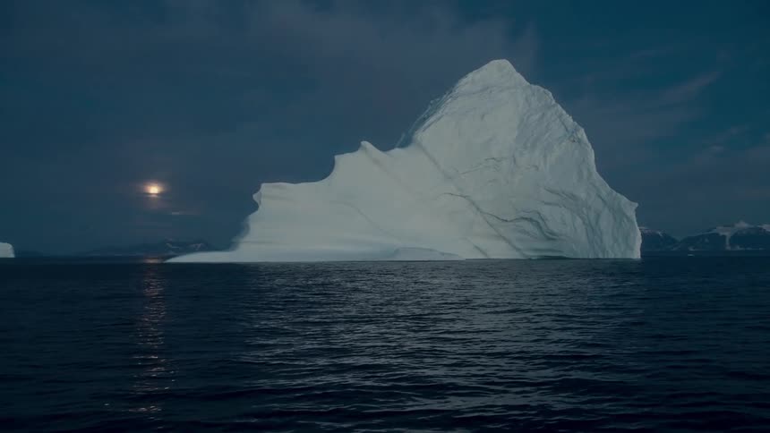A massive iceberg floats in the tranquil Arctic ocean as the full moon rises over the snowy mountains