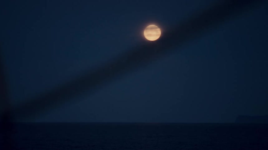 A bright full moon hangs in the dark blue night sky over the tranquil ocean, viewed from a distance