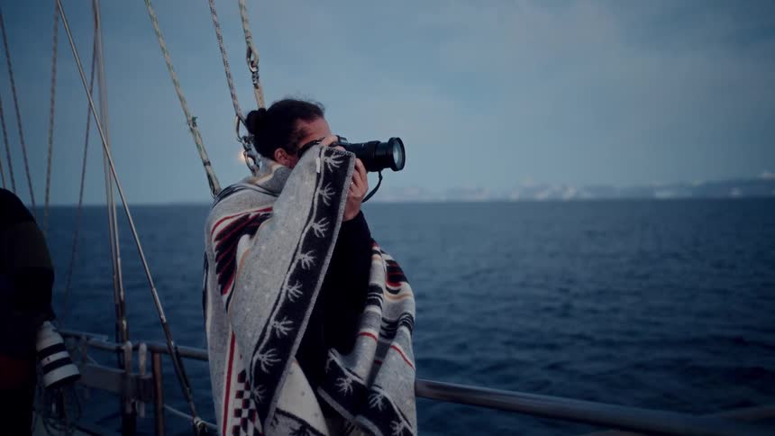 A photographer wrapped in a blanket stands on the deck of a sailboat, capturing images of the vast, cold ocean with snowy mountains on the horizon