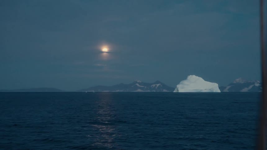 A bright moon hangs in the dusky sky over a calm arctic ocean with a large iceberg and snowy mountains