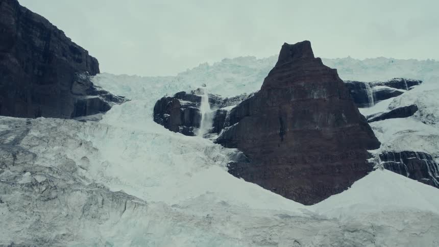 A powerful waterfall of glacial meltwater cascades down a vast, icy mountain range under an overcast sky