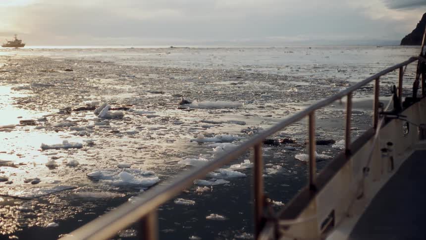 A boat slowly moves through a calm, icy sea filled with floating ice chunks as the sun sets on the horizon