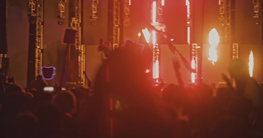 An energetic crowd of silhouetted fans dances and cheers in front of a stage with a spectacular pyrotechnics and light show at a music festival