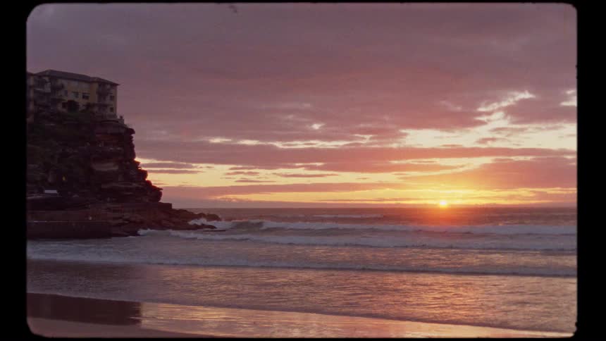 Vintage-style footage of a beautiful sunrise over a calm ocean beach with waves lapping the shore next to a cliffside building