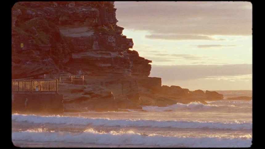 Vintage-style footage shows gentle ocean waves crashing against a rocky cliffside during a serene golden hour