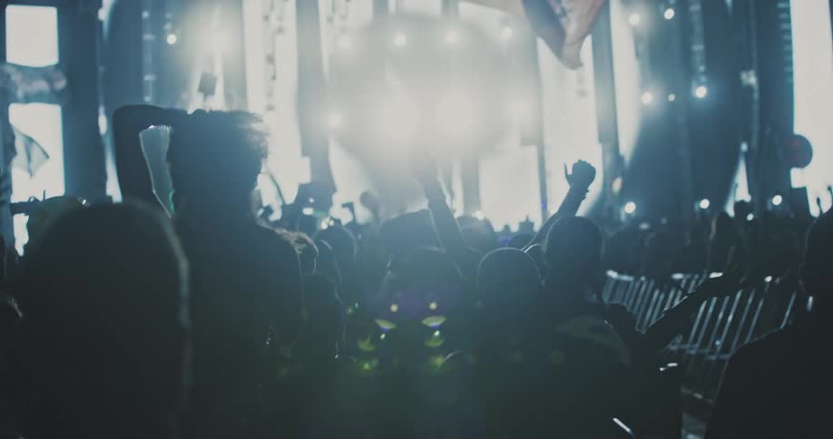 Slow motion view of a silhouetted crowd dancing and headbanging in front of a brightly lit stage at an energetic music festival