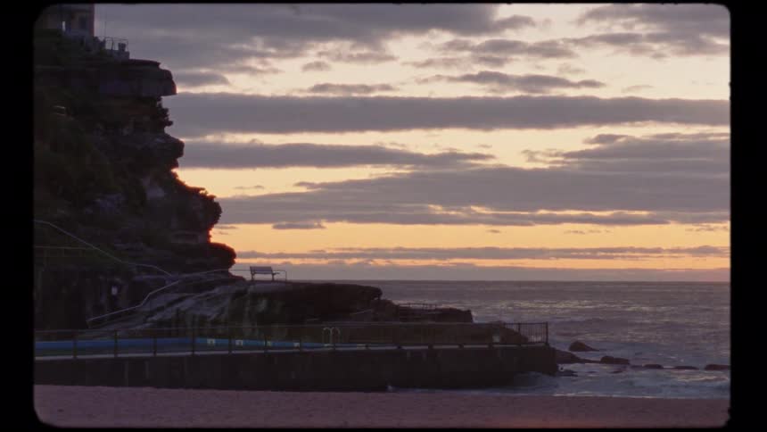 A peaceful, retro-style shot of a rocky cliff and a seaside pool overlooking the ocean during a cloudy sunrise
