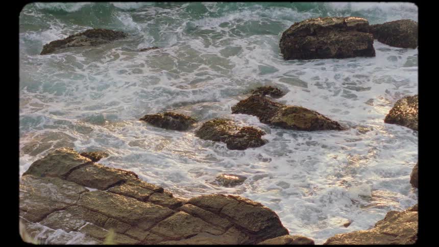 Slow motion vintage footage of ocean waves crashing against a rocky shore at golden hour