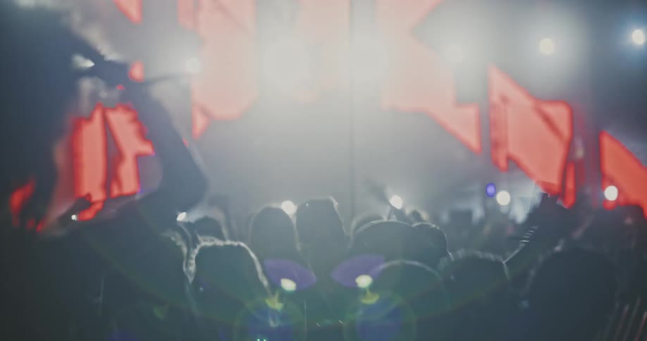 An energetic crowd is silhouetted against a brightly lit stage, dancing and raising their hands at a nighttime concert with a stunning light show