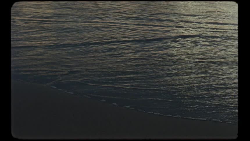 Slow motion vintage footage of gentle ocean waves receding from the sandy shore at dusk