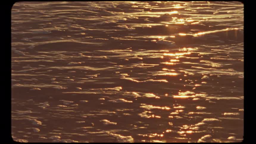 Golden sunlight sparkles on gentle ocean waves washing ashore during a beautiful sunset in vintage-style footage