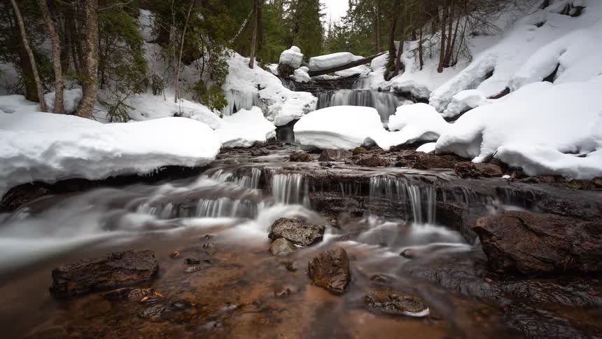 A serene long-exposure timelapse shows a cascading river flowing through a snowy winter forest