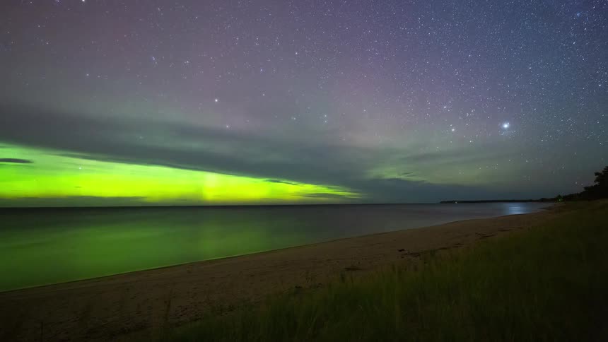 A vibrant timelapse of the green and purple Aurora Borealis dancing in a starry night sky over a calm beach