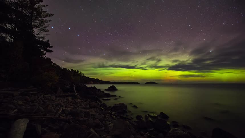 A stunning timelapse captures the vibrant green and purple Aurora Borealis dancing in the starry night sky over a tranquil lake and rocky shoreline