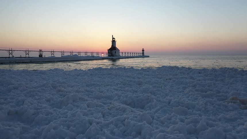 A beautiful sunset casts a warm glow over a frozen lake as a lighthouse stands guard on a snow-covered pier, with icy chunks covering the shoreline