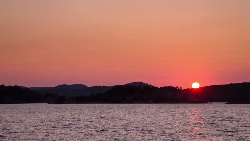 A beautiful time-lapse shows the sun setting behind a silhouetted shoreline, casting a vibrant orange and red glow across the sky and water