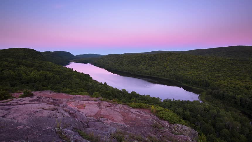 A stunning timelapse from a mountain overlook captures the vibrant pink and purple hues of dusk settling over the tranquil Lake of the Clouds