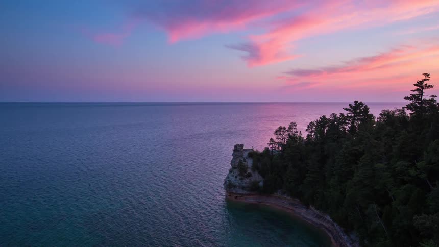 A tranquil view of the rocky coastline of Lake Superior with a vibrant pink and purple sunset reflecting on the calm water